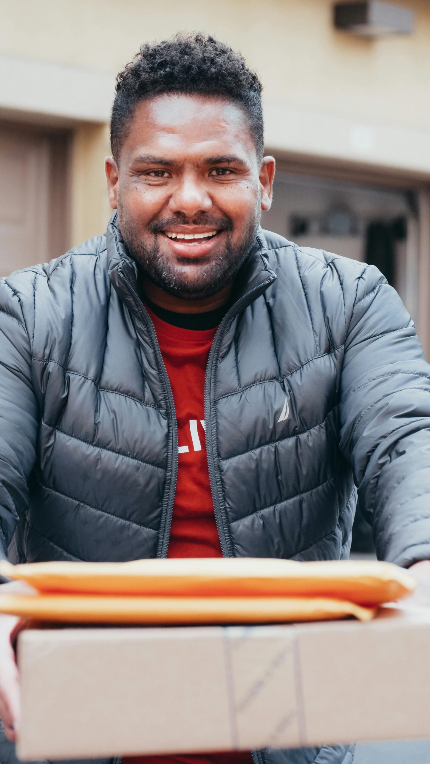 A cheerful deliveryman with a puffer jacket holding envelopes and a parcel outside.